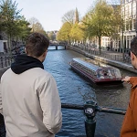 Three guys leaning on a canal bridge railing watching a tour boat pass through an Amsterdam canal