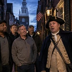 Costumed guide holding a lantern leads a group of men on a nighttime ghost tour through a historic brick alley
