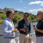 Three middle-aged men toasting with red wine on the deck of a river cruise ship passing a hilltop chateau and vineyards along a French river