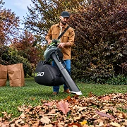 Man using an EGO leaf vacuum and mulcher on a lawn covered in fall leaves, with paper yard bags behind him