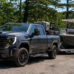 A dark gray GMC Sierra Denali HD pickup towing a center-console fishing boat on a tandem trailer at a tree-lined northern Michigan lake boat ramp