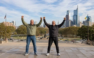 Two men at the top of the Philadelphia Museum of Art steps with their arms raised, the Benjamin Franklin Parkway and the Center City skyline behind them